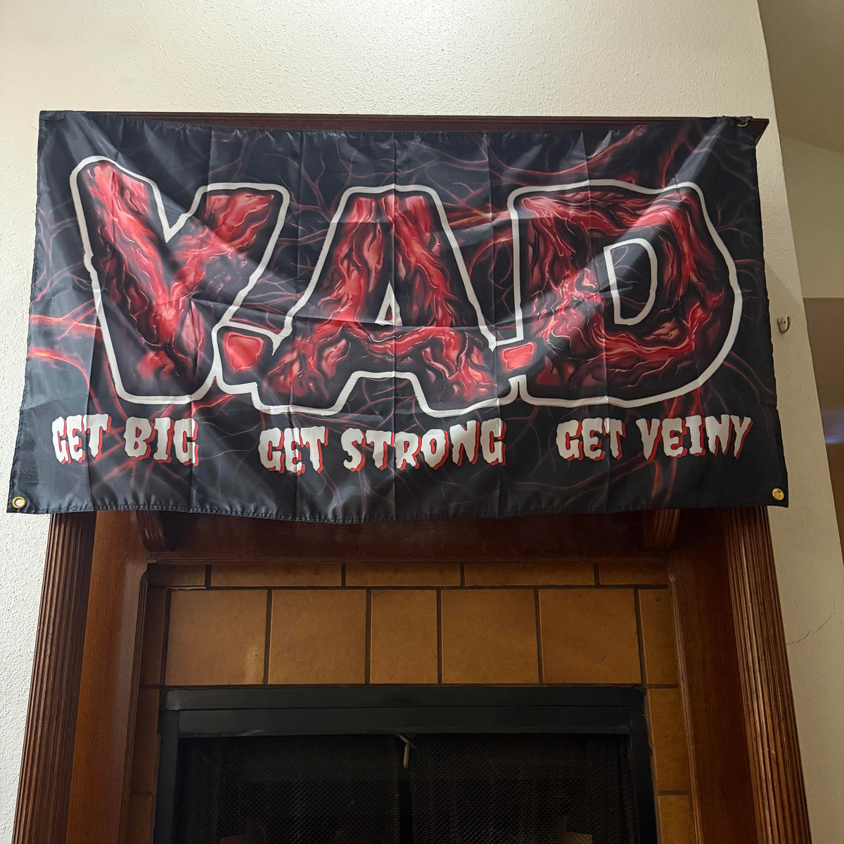 A black banner with the word 'VAD' in large red marbled letters outlined in white, with the phrases 'GET BIG GET STRONG GET VEINY' below in red and white, hanging above a fireplace.