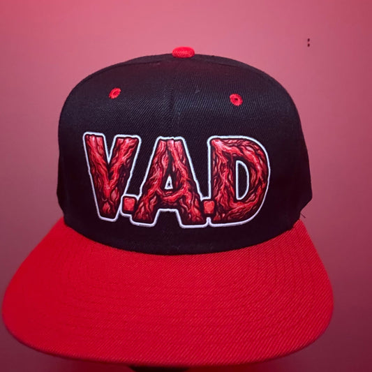 A black and red snapback cap featuring the word 'VAD' embroidered on the front panel in a red textured font with a white outline, set against a pinkish-red background.