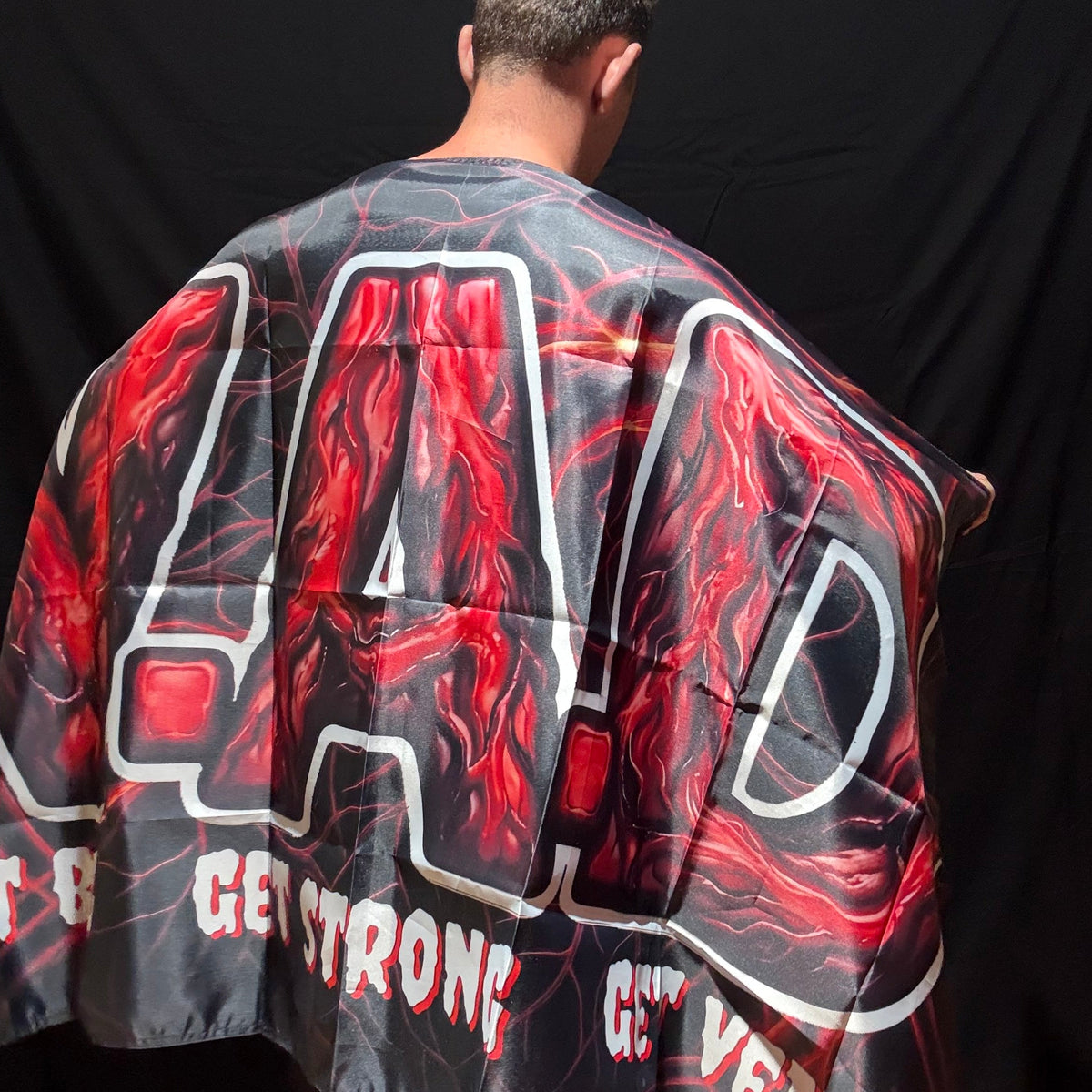 A man viewed from behind wearing a black and red V.A.D flag with large white outlined letters and text, standing against a black backdrop.