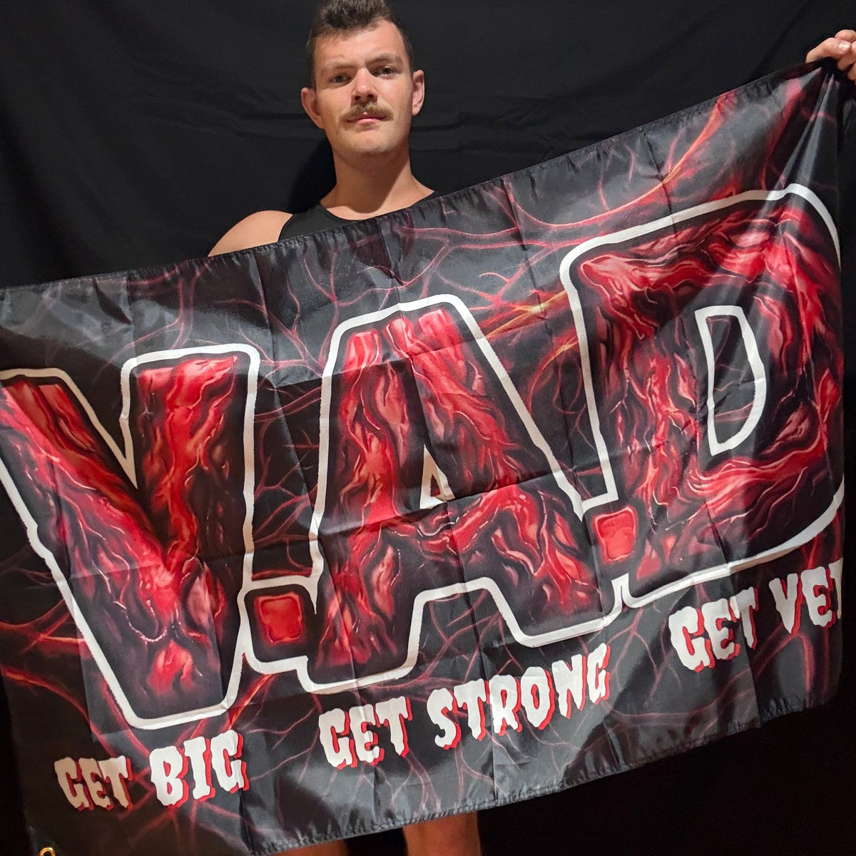 A man holding up a black banner with a red and white graphic design featuring the text 'VAD GET BIG GET STRONG GET VEINY' in stylized lettering.