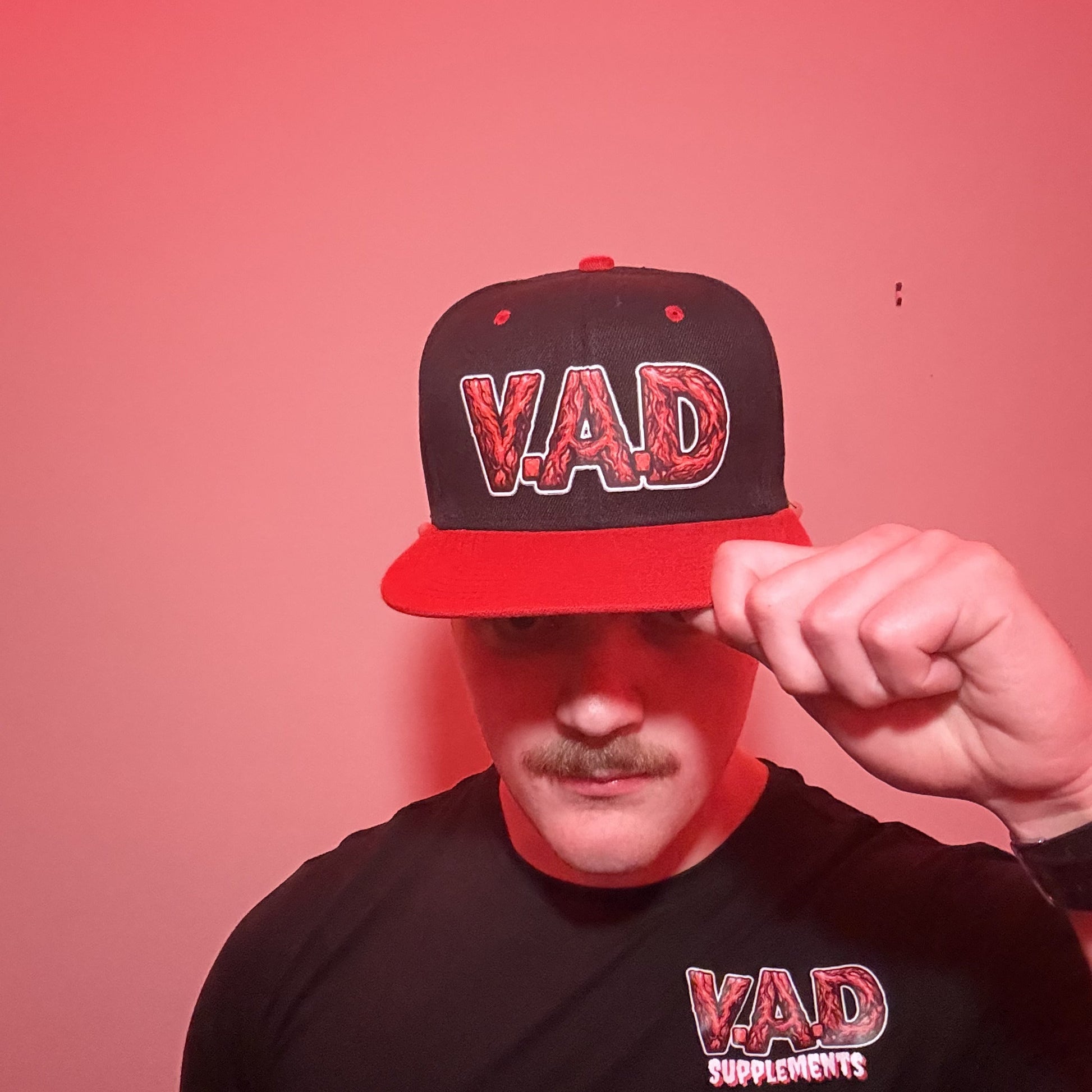 A man wearing a black and red snapback cap with 'VAD' embroidered on the front, holding the brim over his eye, and wearing a matching black t-shirt with a red and white 'VAD SUPPLEMENTS' logo, set against a red-lit background.