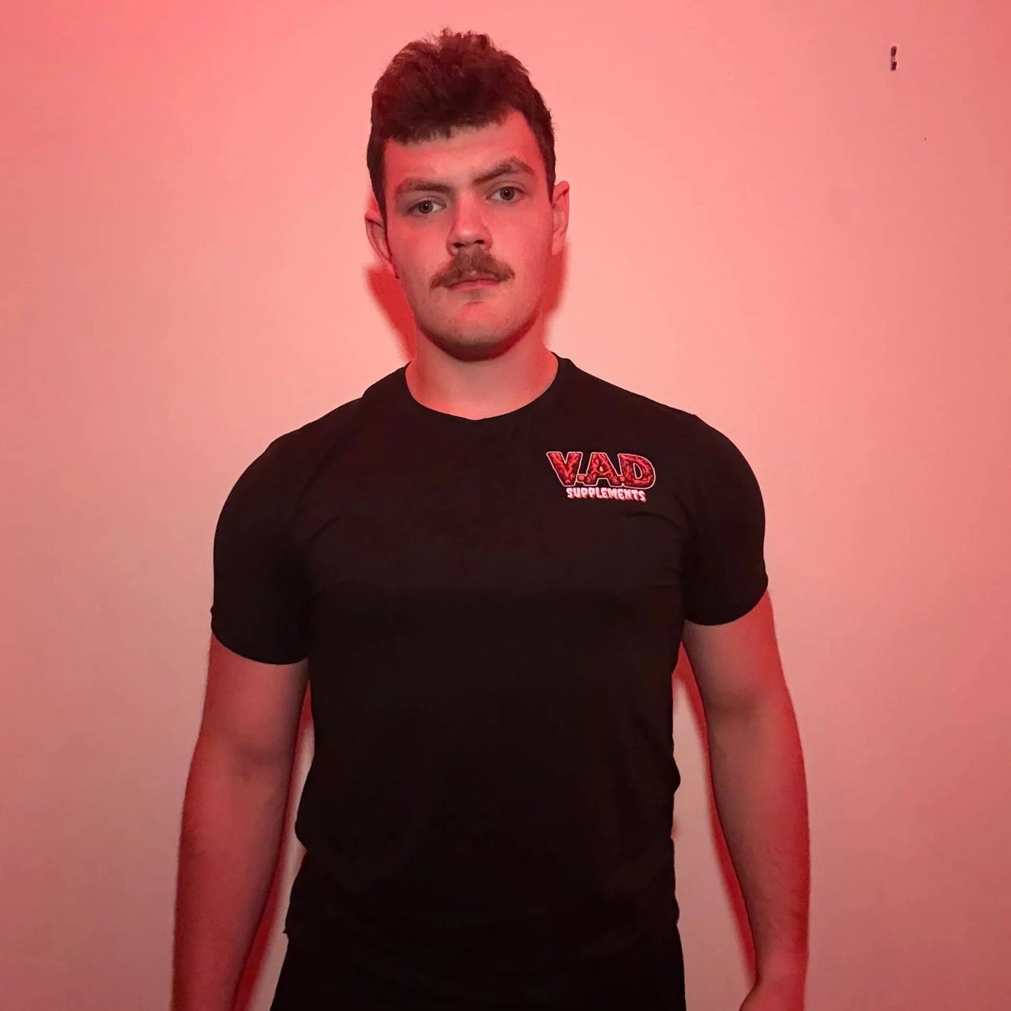 A man with a mustache wearing a black short-sleeve t-shirt with a red and white 'VAD Supplements' logo on the left chest, standing against a pinkish-red lit wall.