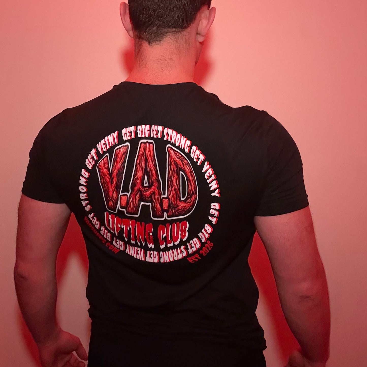A man viewed from behind wearing a black t-shirt with a large red and white graphic on the back that reads 'V.A.D lifting club'