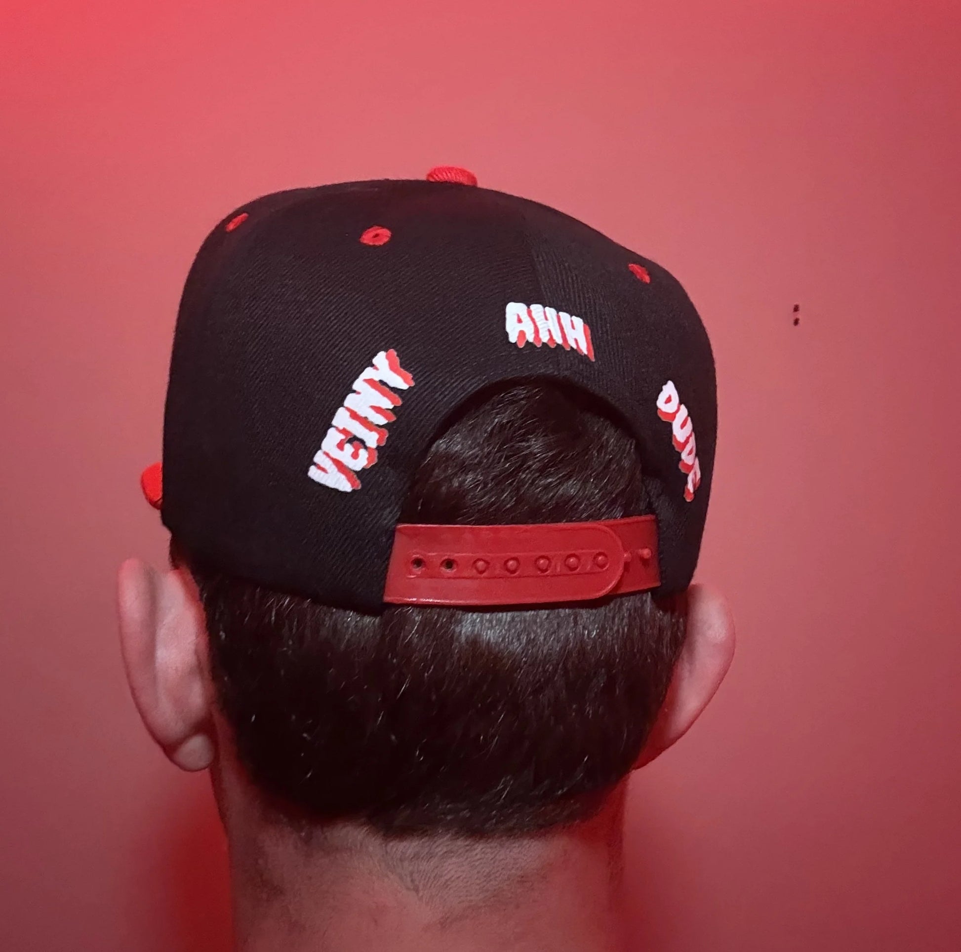 A black baseball cap with red and white embroidered text, shown from the back on a person's head against a red background.