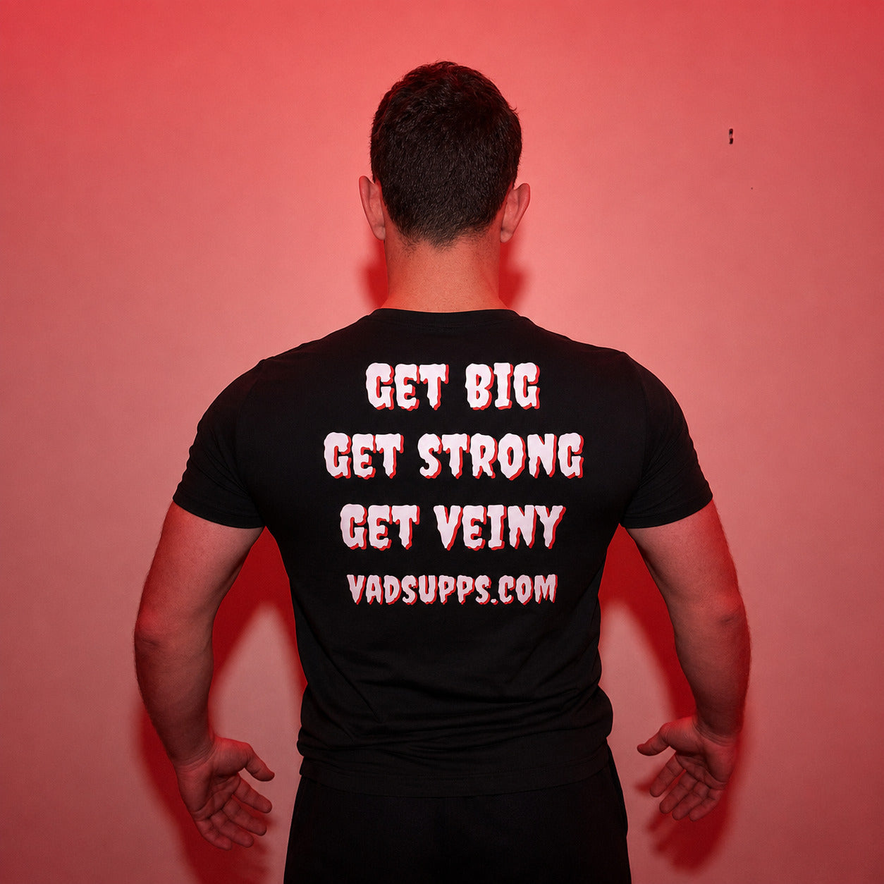 A man viewed from behind wearing a black t-shirt with the phrase 'GET BIG GET STRONG GET VEINY' and 'VADSUPPS.COM' printed in white with a red outline on the back, set against a red-lit background.